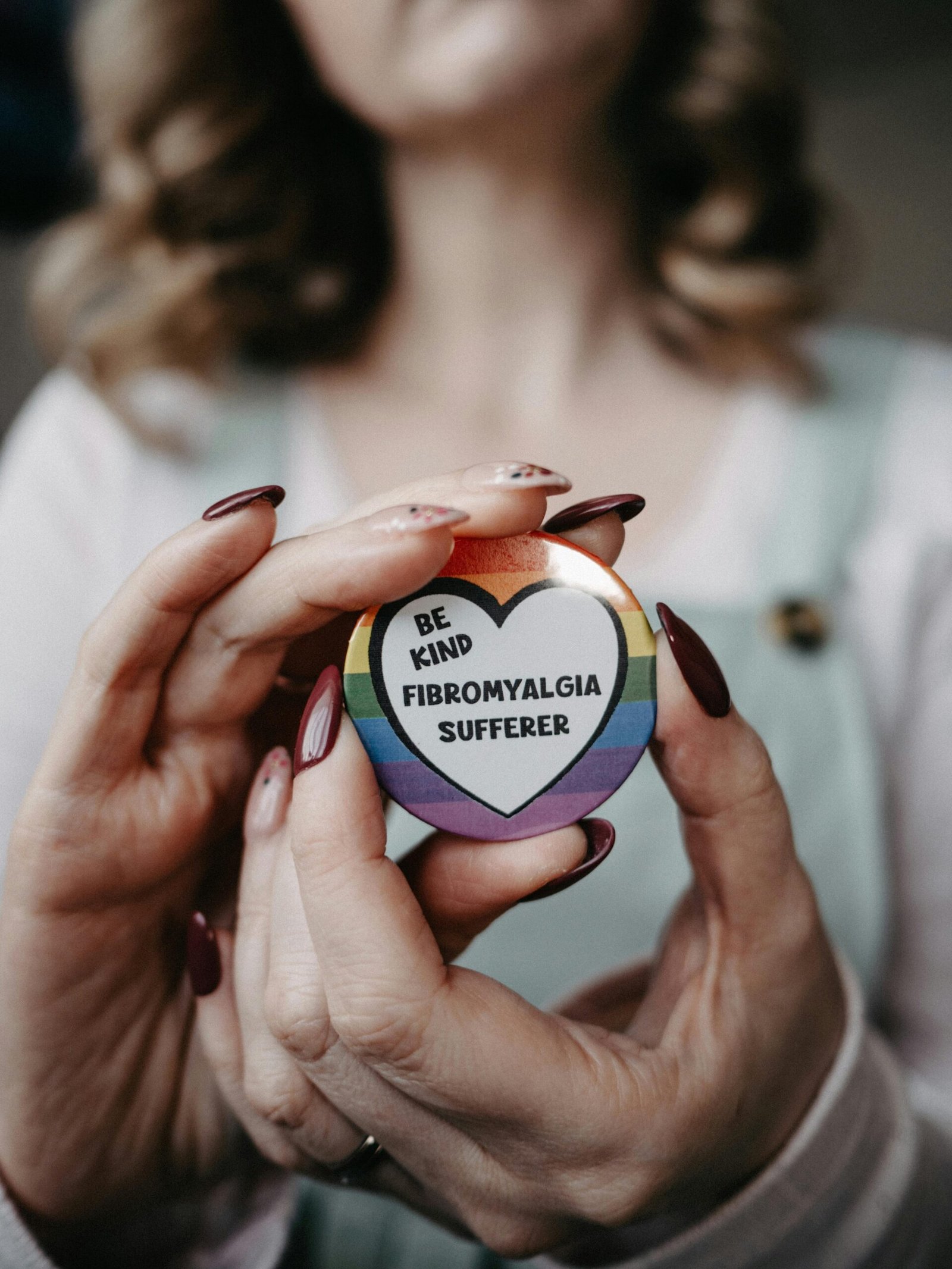 Hands holding a 'Be Kind - Fibromyalgia Sufferer' badge promoting awareness.