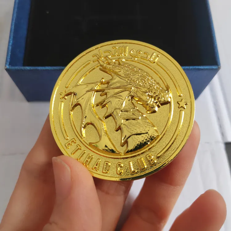 A shiny gold commemorative coin held by a hand. The center features a detailed eagle emblem with "ETIHAD CLUB" inscribed at the bottom and Arabic calligraphy at the top.
